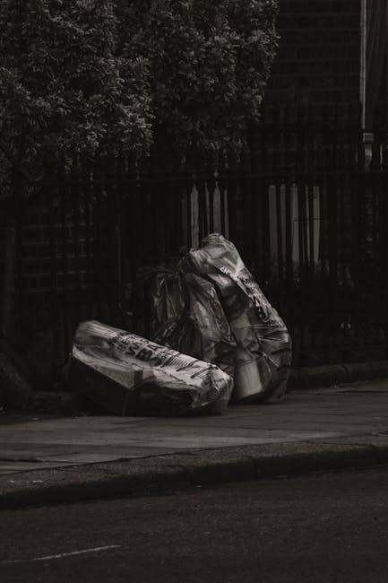 Three large black plastic trash bags filled with waste are placed on a paved sidewalk near a black metal fence. The bags are crumpled and slightly torn, revealing some contents inside. They are positi