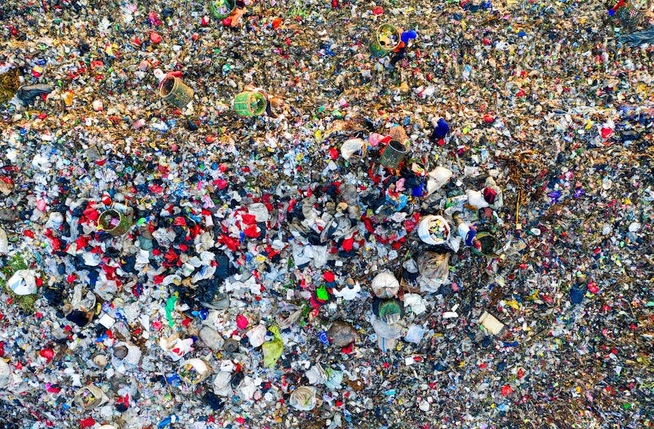 An aerial view of a large landfill site filled with a mixture of waste materials, including plastic bottles, paper, and general household rubbish scattered across the uneven, gravelly terrain. Two yel